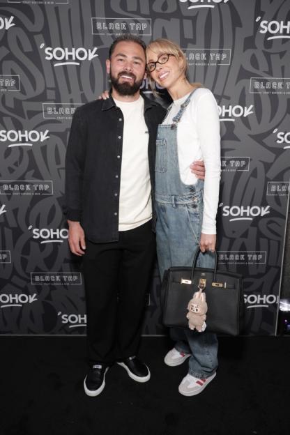 A man with a beard and a woman in overalls standing against a Black Tap Soho background.