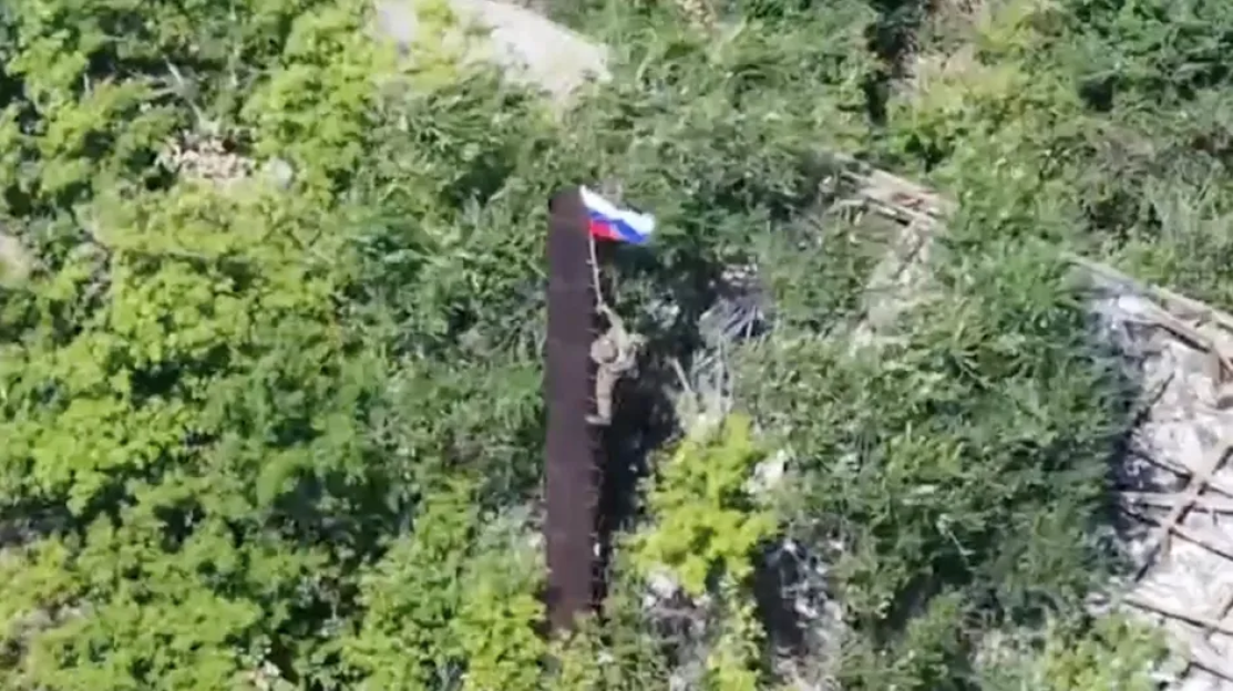 Aerial view of a soldier raising a Russian flag on a tall pole.