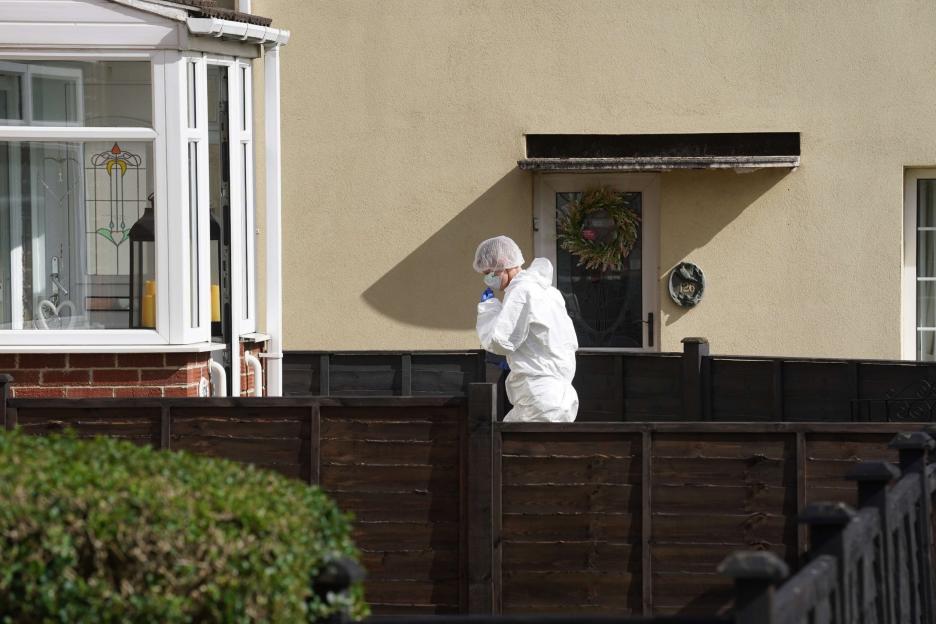 Crime scene officer in a white forensic suit and hairnet walks behind a fence.
