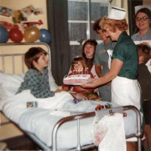 A nurse presents a birthday cake to a young boy in a hospital bed.