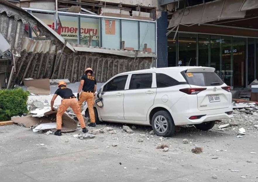 Two fire officers clearing debris from outside a shopping mall after an earthquake, with a white car next to them.