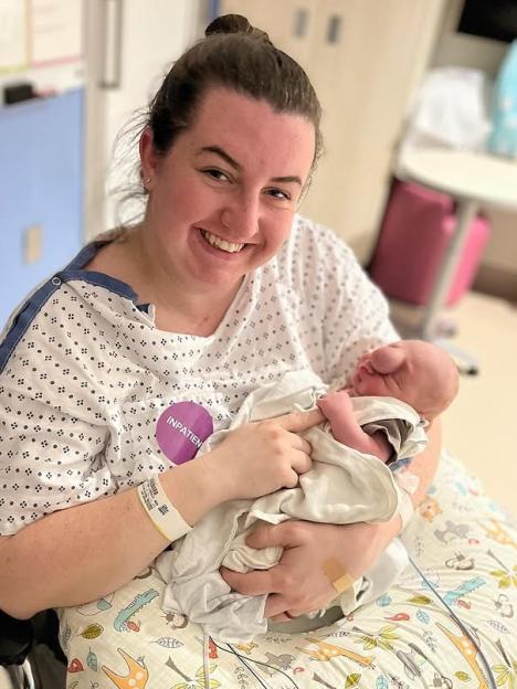 Hannah Sachs smiles while holding her newborn baby, Liam, wrapped in a blanket.