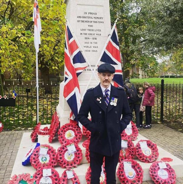 Man in military-style coat standing by a war memorial with wreaths.