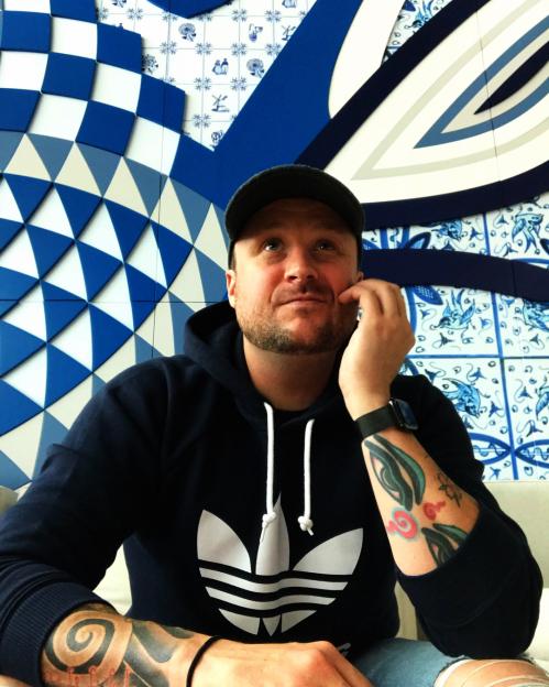 Brooklyn Earick in a black Adidas hoodie, cap, and ripped jeans, with a patterned wall behind him.