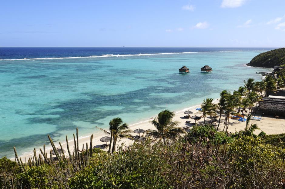 View of Raffles Resort on Canouan Island, Saint Vincent, with two bungalows in the turquoise ocean, a sandy beach with thatched umbrellas, and palm trees.