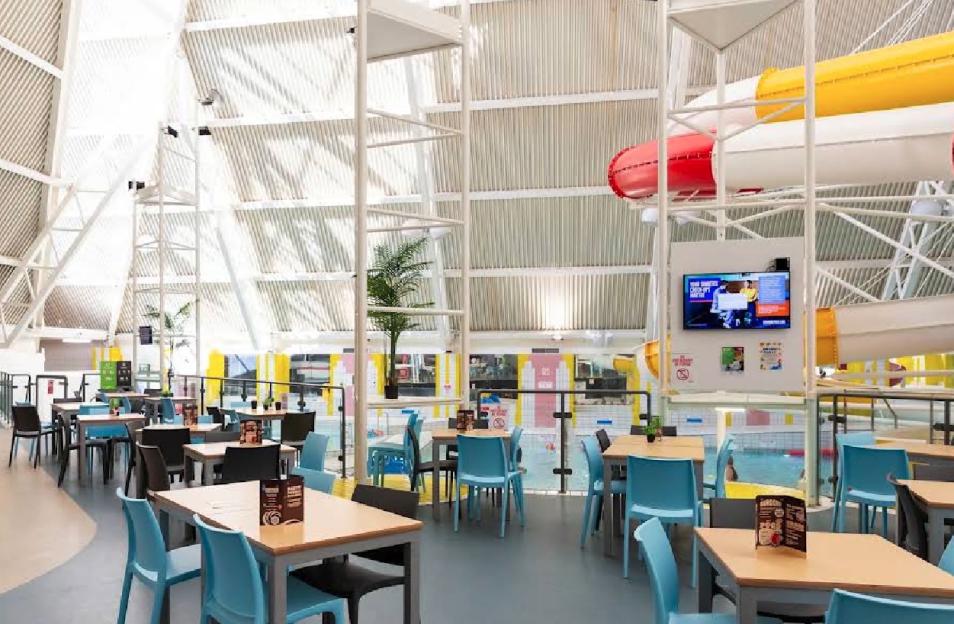 Oasis Beach Swimming Pool indoor cafe seating area with tables and chairs next to the swimming pool, with water slides in the background.