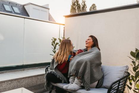 Two women wrapped in a blanket on a patio, enjoying drinks together.