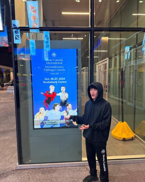 A man in a black hoodie and sweatpants gesturing towards a digital poster for Skate Canada International.