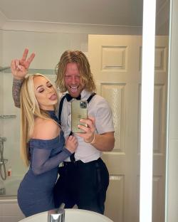 Selfie of a couple in a bathroom; the woman is wearing a blue dress and making a peace sign, and the man is wearing a white shirt and black pants.