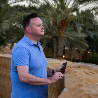 Man in blue shirt holding sunglasses, standing outdoors.