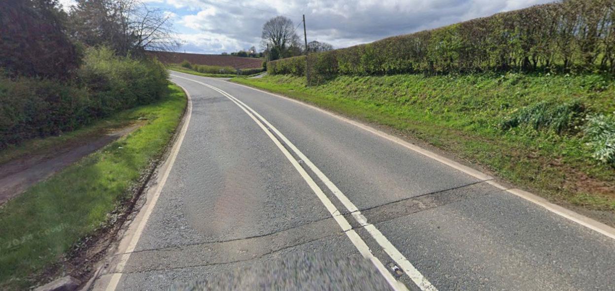 An empty, winding road surrounded by green hedges and fields under a cloudy sky.