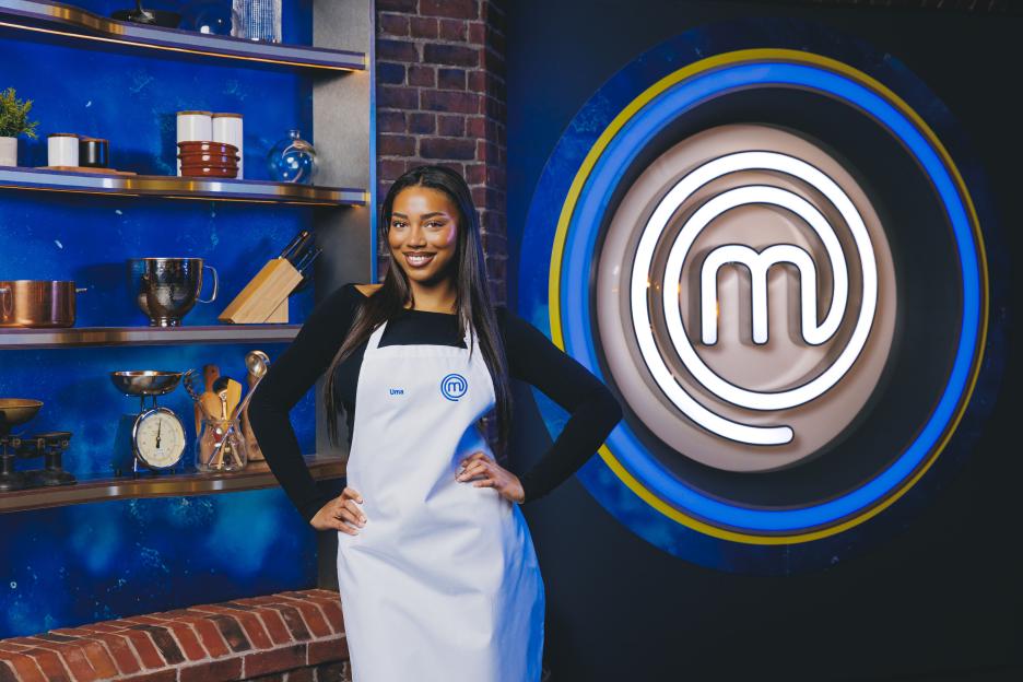 Uma, a contestant on Celebrity MasterChef, stands in a kitchen set wearing a white apron.