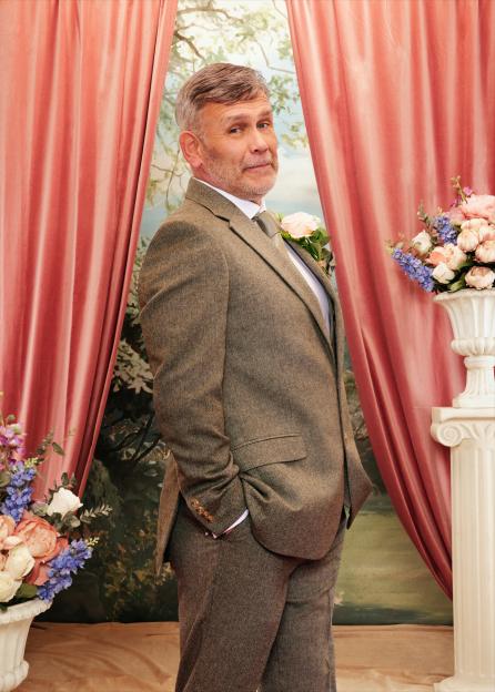 Paul wearing a brown tweed suit with a peach-colored rose boutonnière, looking over his shoulder.