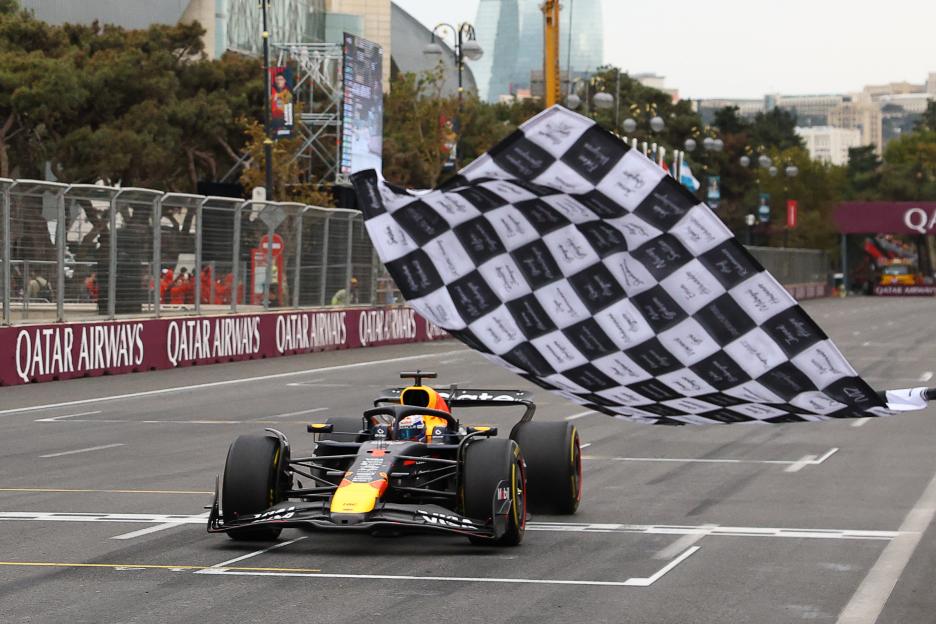 Max Verstappen in a Red Bull Racing car crosses the finish line at the Formula One Azerbaijan Grand Prix, with a checkered flag waving above.