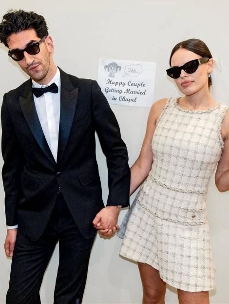 A man and woman, both wearing sunglasses, holding hands in front of a sign that reads "Happy Couple Getting Married in Chapel."