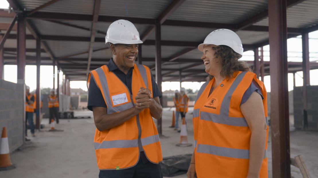Two people in hard hats and high-visibility vests laugh together at a construction site.