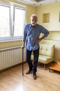Older man with physical disabilities standing indoors with a cane.