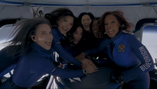Six women in blue flight suits huddle together in a spacecraft.