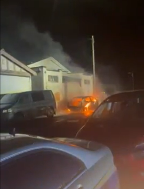 A car on fire at night on a residential street, with smoke rising into the dark sky.