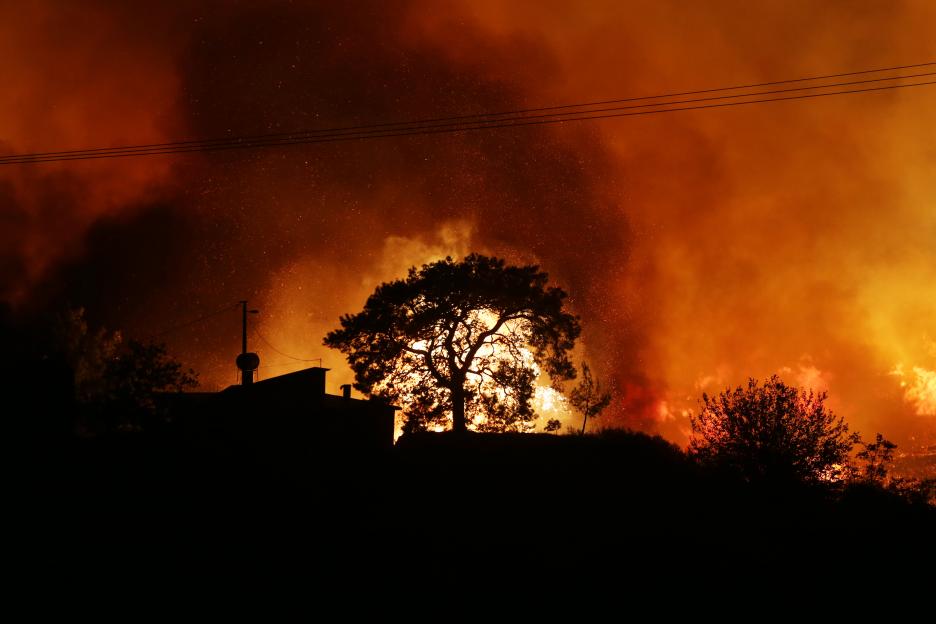 Silhouetted trees and buildings against an orange and red sky filled with smoke and sparks from a wildfire.