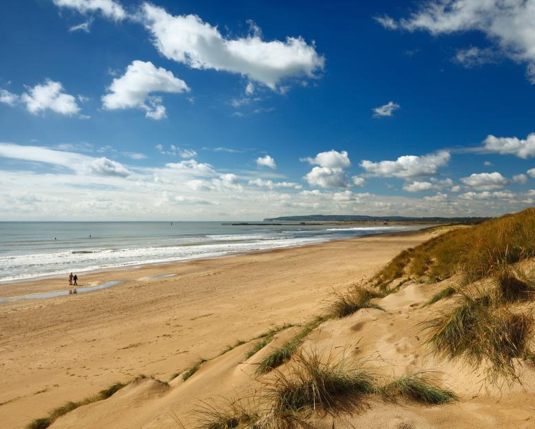 Camber Sands beach with two people walking near the water and grassy dunes.