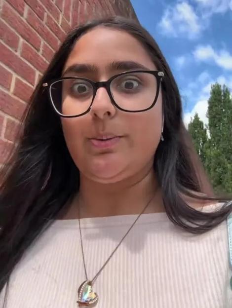 A woman wearing glasses looks at the camera with wide eyes and open mouth, standing next to a brick wall under a blue sky with some text overlay.