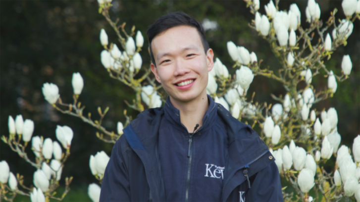 Charles Shi from Kew Gardens in a blue jacket, smiling in front of white flowers.