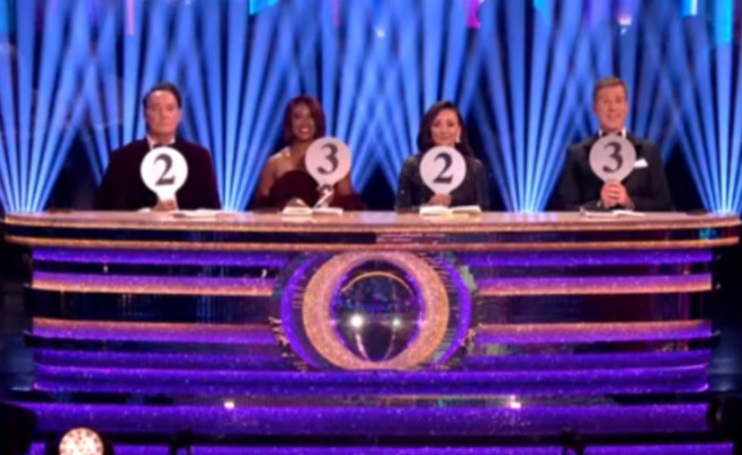 Four judges at a desk holding up score paddles, with two showing a 2 and two showing a 3.