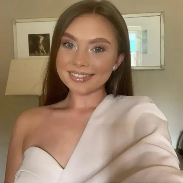 Headshot of a smiling young woman with long brown hair, wearing a light-colored off-the-shoulder top.