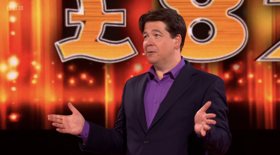 Man in suit presenting in front of a game show backdrop.