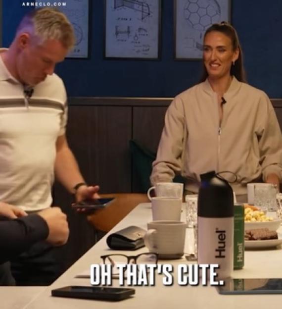 Two people sitting at a table with drinks, with a man bending over to look at something on his phone.