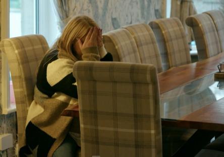 A woman sits at a table, head in her hands, appearing upset.