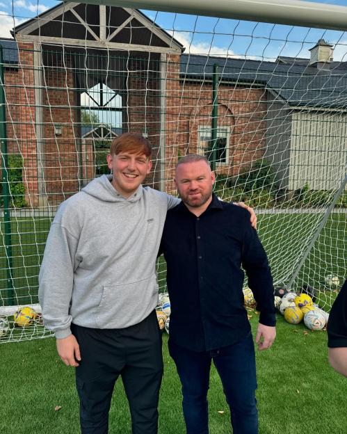 Wayne Rooney standing next to a younger man, with the younger man's arm around Rooney, in front of a soccer goal.
