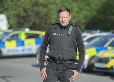 Police officer in tactical vest standing in front of police cars.