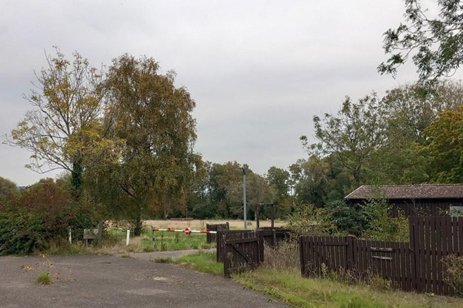 A path leading to a closed barrier gate with a "do not enter" sign, a fence, and a building on the right, surrounded by trees and overgrown vegetation.