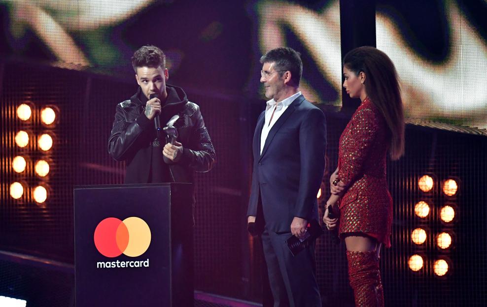 Liam Payne speaking into a microphone while Simon Cowell and Nicole Scherzinger stand beside him at the Brit Awards.
