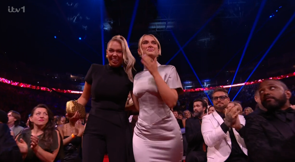 Two women walking and smiling, surrounded by an audience applauding.