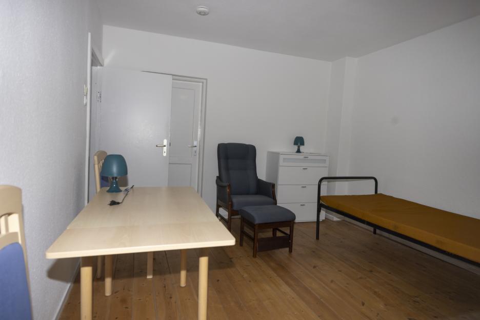 A plain room with white walls, a light wooden table, dark blue chair and footrest, a white chest of drawers, and a narrow bed with a mustard-yellow mattress.