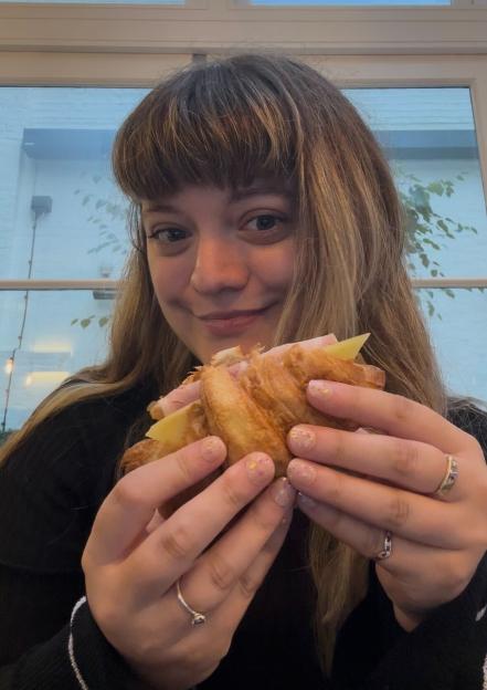 A smiling woman holding a croissant with ham and cheese.