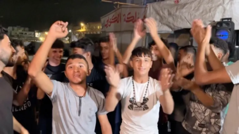 A group of men and boys celebrating and clapping, with Arabic writing on a sign in the background.