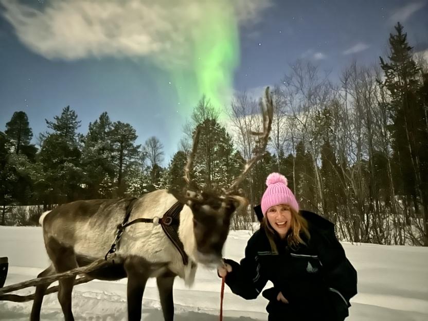A person wearing a pink hat holding the reins of a reindeer, with the Northern Lights visible in the background.