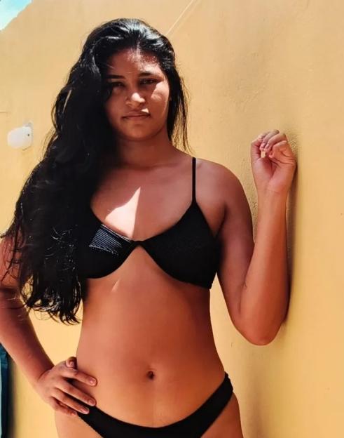 Woman in black bikini leaning against a yellow wall.