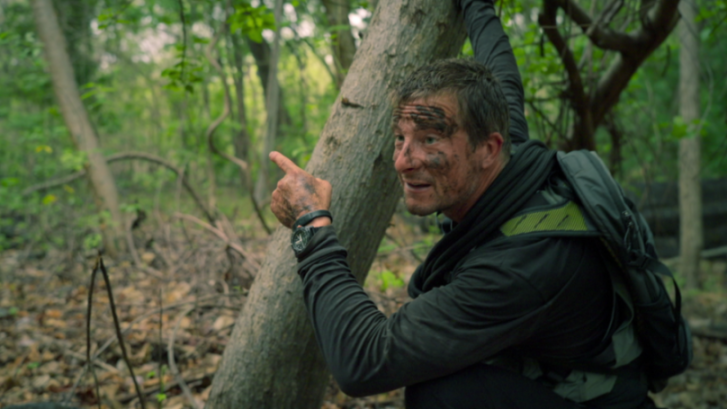 Bear Grylls in a forest, his face and hand covered in dirt, pointing to the left.