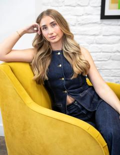 Portrait of Lily Phillips in a denim vest and jeans, sitting in a yellow armchair.