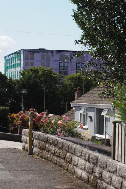 Abandoned purple student accommodation block in Penryn, Cornwall, with boarded-up windows and green netting, towering over houses with overgrown gardens.