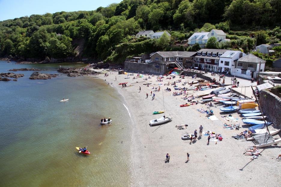 Aerial view of Cawsand beach in Plymouth, England, with people sunbathing and enjoying water activities.