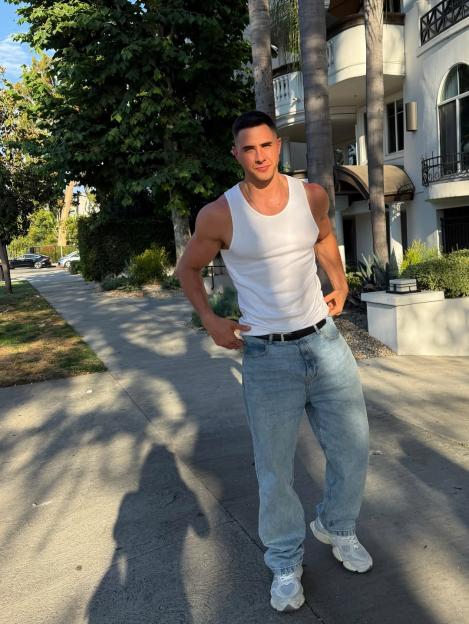 Man in white tank top and jeans standing on a sidewalk.