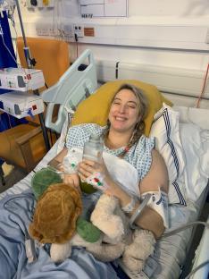 Woman in hospital bed holding a drink and stuffed animals.