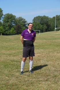 Man standing on a grass field wearing a purple shirt, black shorts, and soccer socks.