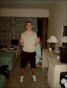 A young man standing in a hotel room, making a hand gesture.